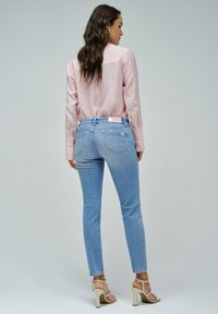 Camisa de manga larga rosa clara, jeans ajustados azul claro, vista posterior, con un pequeño detalle de bolsillo trasero, combinada con sandalias de tacón beige.