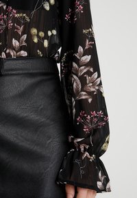 Svart blommig blus med genomskinligt tyg, försedd med rosa och gröna botaniska mönster. Ärmarna är utsvängda med delikata volanger. Tillsammans med en svart läderkjol.