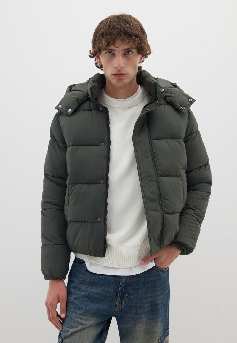 Giubbotto imbottito verde scuro con zip e bottoni a pressione, dotato di cappuccio e texture trapuntata, indossato sopra un maglione bianco e jeans blu.