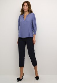 Helle lila Bluse mit langen Ärmeln, V-Ausschnitt und Rüschenmanschetten, kombiniert mit taillierten navyblauen Hosen und schwarzen Pumps mit spitzem Schuhwerk.