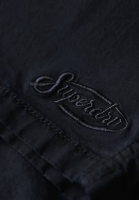 Tela negra con un logo bordado de "Superdry" en hilo a juego. La textura es suave y se pueden ver las costuras a lo largo del borde.