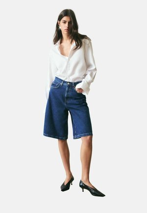 Kobieta ubrana w oversize'ową białą koszulę na guziki, długie bermudy z dżinsu, czarne czółenka na niskim obcasie oraz srebrne kolczyki.