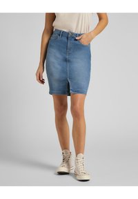 Jupe crayon en jean bleu clair avec une fente à l'avant, taille haute, cinq poches, et une fermeture classique à bouton et fermoir éclair. Portée avec des baskets montantes beiges.