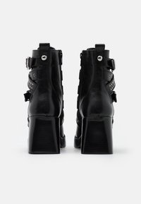 Botas de cuero negras hasta el tobillo con tacones gruesos, que presentan detalles de hebillas y tachuelas en los laterales. Textura suave con acabado brillante.