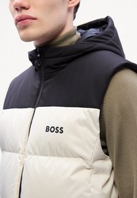 Lettvektet hettegenservest i krem og svart, med quiltet tekstur. "BOSS"-logoen er vist i svart foran.