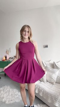 Uska haljina bez rukava u tamno magentnoj boji s plisiranim suknjom, izrađena od glatkog materijala. Dodaci uključuju naušnice na obruče i ležerne crne patike.