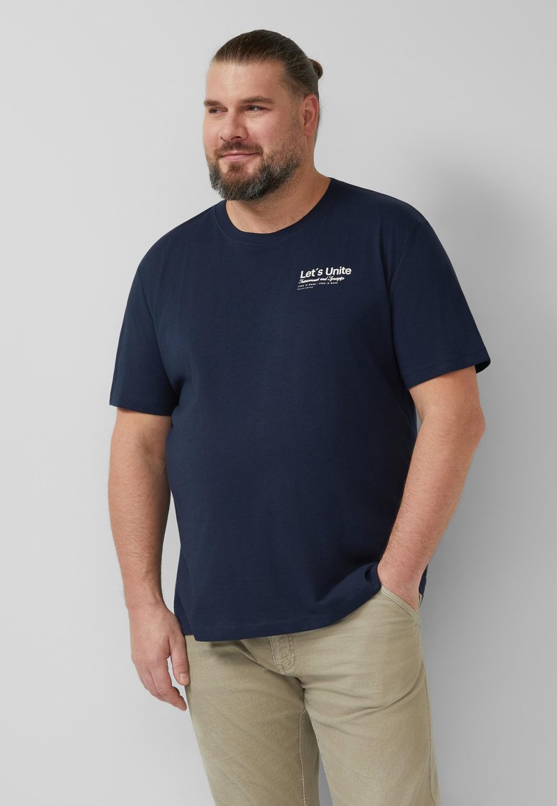 Homme barbu portant un T-shirt bleu marine avec l'inscription "Let's Unite" et un pantalon beige, debout avec une main dans la poche sur un fond clair uni.