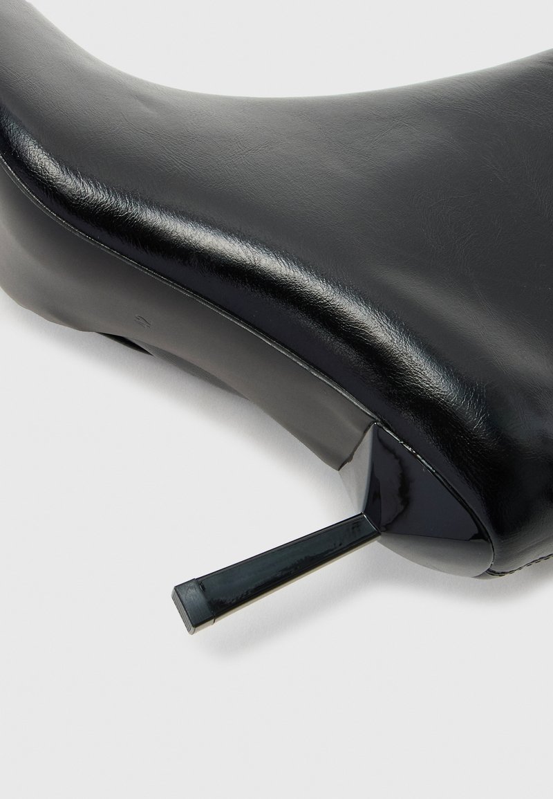 Primo piano di una scarpa nera in pelle con tacco alto, incentrato sul tacco appuntito e sulla texture liscia della parte superiore.