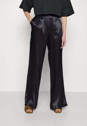 Vrouw draagt zwart satijnen broek met hoge taille en wijd uitlopende pijpen, met hand in de zak, gecombineerd met gouden open-toe haksandalen tegen een witte achtergrond.