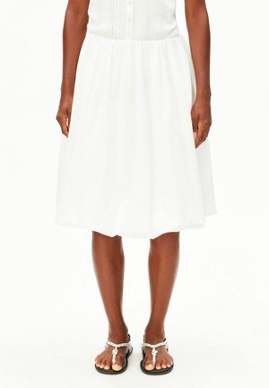 Frau trägt einen weißen knielangen Rock mit Knöpfen und geflochtenen Riemchensandalen, steht vor einem schlichten weißen Hintergrund.