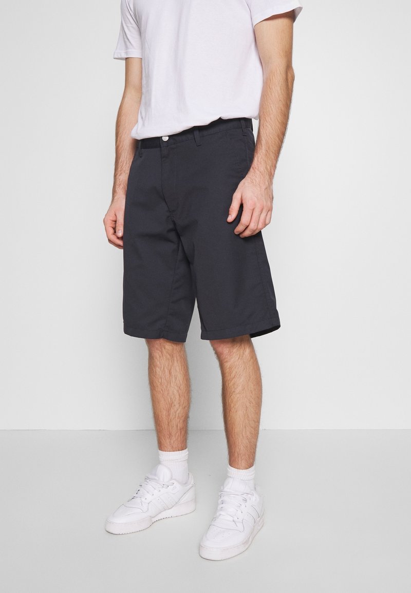 Mörkblå shorts i bomullsmaterial med en blandning av tyger, med rak front, bälteshällor och en lätt glans. Bärs med vita sportskor.