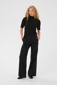 Žena s kučeravými blond vlasmi, oblečená v čiernom krátkom roláku, čiernych širokých nohaviciach a čiernych balerínach, stojí s rukami v vreckách.