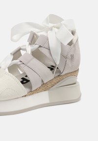 Baskets gris clair avec lacets ruban, bout en blanc texturé, sangles élastiques et semelle compensée de style espadrille. Accents lisses partout.