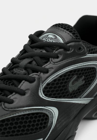 Chaussure de sport noire avec un design en mesh, des accents en cuir lisse, des lacets plats et un logo discret. Semelle en caoutchouc durable avec une finition texturée.