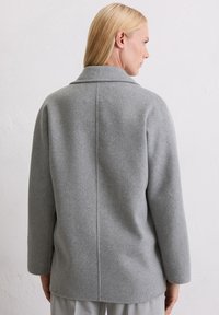 Donna con capelli biondi che indossa un cappotto di lana grigio chiaro, in piedi di spalle contro uno sfondo di parete chiara e uniforme.