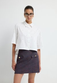 Witte, geknipte button-up shirt met verspreide versieringen, gecombineerd met een textuur minirok in marineblauw met gouden accenten op de zakken.