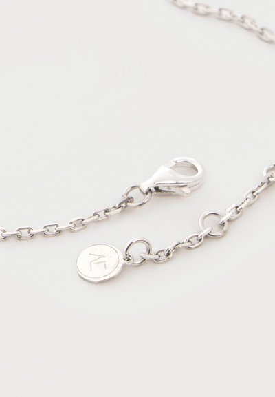Ασημένιο κολιέ αλυσίδα με κούμπωμα καβουράκι και μια μικρή στρογγυλή ετικέτα χαραγμένη με ένα στυλιζαρισμένο γράμμα σε φωτεινό φόντο.
