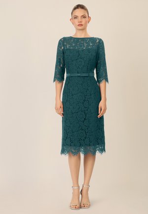 Tefarbene Spitzenkleid mit Dreiviertelärmeln, einem runden Ausschnitt und einem gewellten Saum, ausgestattet mit einem passenden Stoffgürtel. Silberne Riemchen-Absätze.