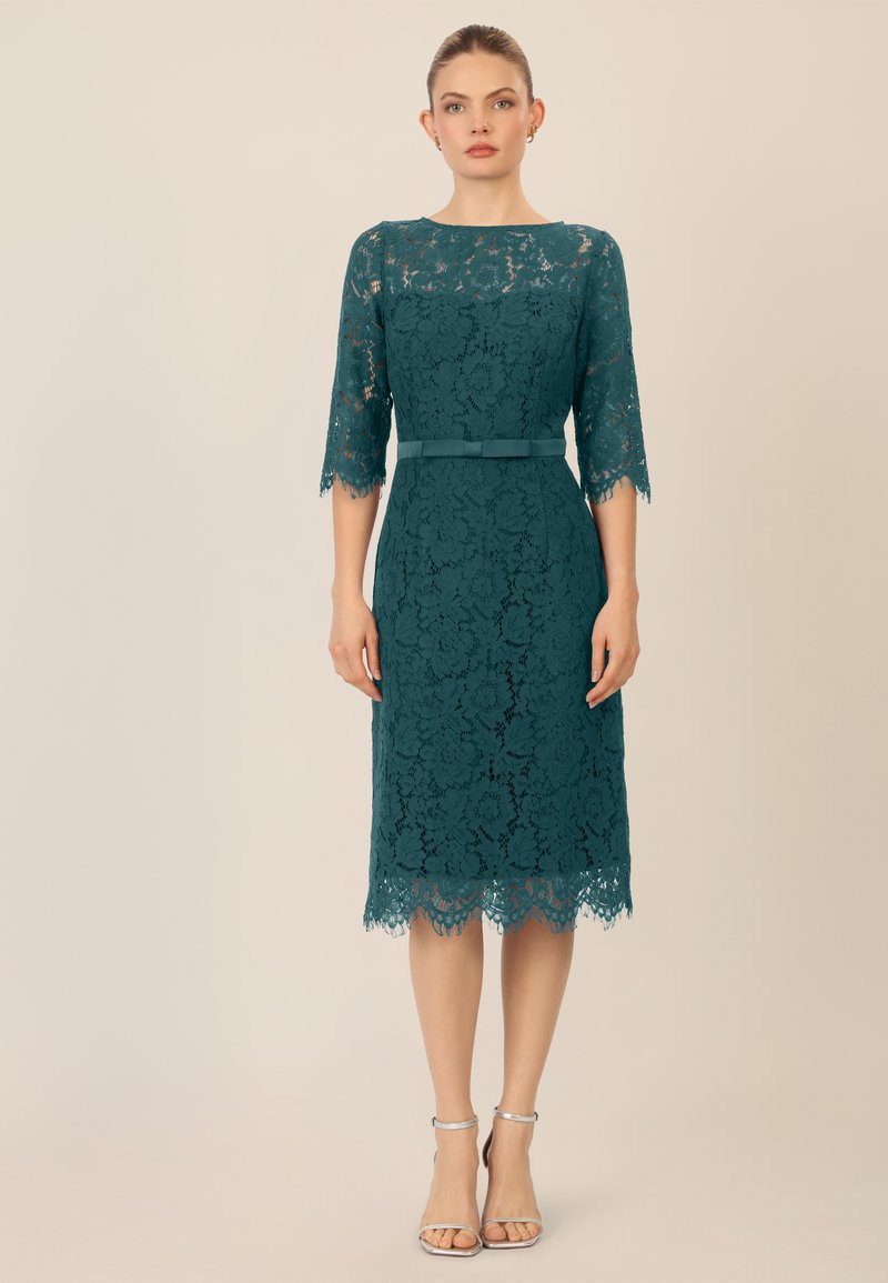 Abito in pizzo teal con maniche a tre quarti, scollatura tonda e orlo ondulato, caratterizzato da una cintura coordinata in tessuto. Tacchi a strappi argentati.