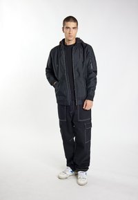 Sort bomberjakke med lynlåslommer, parret med løse cargo-bukser med hvid syning. Modellen bærer hvide sneakers med blå accenter.
