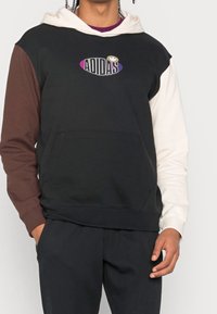 Mikina s kapucí s černým tělem a hnědými a krémovými rukávy. Na hrudi je logo Adidas ve fialové a bílé barvě a přední kapsa.