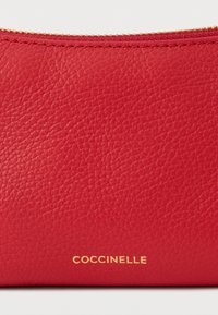 Crvena kožna torbica s teksturiranom površinom, s zlatnim logotipom "Coccinelle" na dnu i zatvaranjem na patentnu zatvarač duž gornjeg ruba.