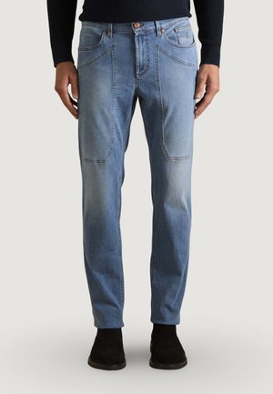 Hombre con vaqueros de mezclilla azul claro con grandes bolsillos frontales, camiseta ajustada negra y botines negros, de pie frente a un fondo blanco.