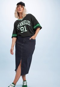 Camisa negra de gran tamaño con "Los Ángeles 91" en letras blancas y verdes; combinada con una falda maxi de mezclilla oscura con una abertura frontal. Zapatillas verdes.