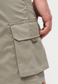 Κοντινό πλάνο ενός χεριού που αγγίζει το καπάκι μιας μπεζ τσέπης τύπου cargo σε σορτς από ελαφρύ ύφασμα.