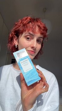 Personne aux cheveux roux courts tenant une boîte bleue de produit de soin de la peau étiquetée « Centella » près du visage, portant une chemise blanche dans une pièce lumineuse.