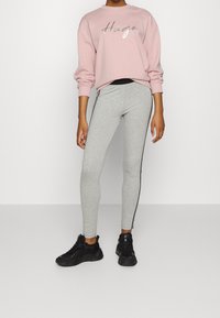 Hellrosa Sweatshirt mit "Hugo" in silberner Schreibschrift, kombiniert mit grauen Leggings mit schwarzen Seitestreifen. Getragen mit schwarzen Turnschuhen.