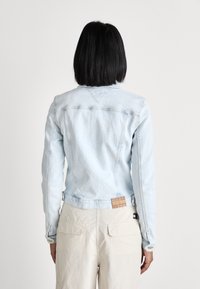 Light blue denim jacket with visible stitching, a cropped design, and two buttons at the waist. Features a brand tag on the back.