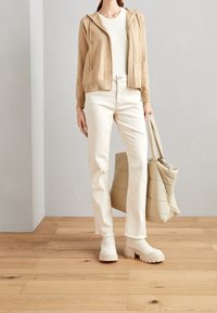 Beżowa bluza z zamkiem, biała koszulka i off-white jeansy dzwony. Masywne beżowe botki i pikowana beżowa torba na ramię. Tło z jasnego drewna.