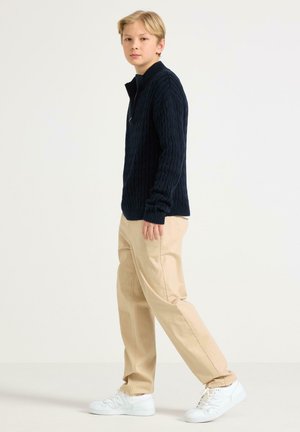 Lichtblond jong persoon staand, gekleed in een marineblauwe kabelgebreide trui, beige broek en witte sneakers tegen een effen lichte achtergrond.