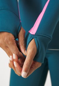 Türkises Sportoberteil mit pinken Akzenten, das über Daumenlöcher in den Bündchen verfügt. Der Stoff wirkt glatt und elastisch, ideal für Sportbekleidung.