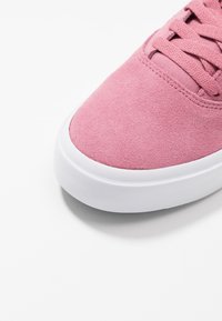 Gros plan d’un bout de chaussure de sport en daim rose avec des lacets assortis et une semelle en caoutchouc blanche sur un fond blanc uni.