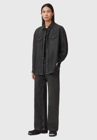 Czarna koszula dżinsowa z dwoma kieszeniami na piersi i spiczastym kołnierzykiem, w połączeniu z szerokimi czarnymi dżinsami. Model stoi, ubrany w czarne botki.