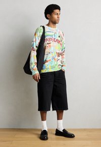 Πολύχρωμο πλεκτό πουλόβερ με γραφικά και κείμενο, συνδυασμένο με μαύρο σορτς, λευκές κάλτσες και μαύρα μοκασίνια. Κρατάει μια μαύρη τσάντα.