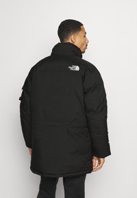 Zwarte geïsoleerde pufferjack met hoge kraag, voorzien van het The North Face-logo op de bovenrug en een gladde, iets glanzende textuur.