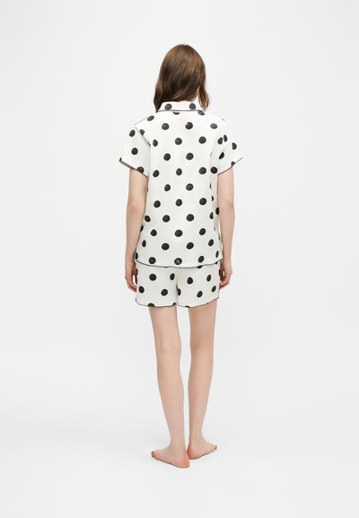 Femme debout, pieds nus, de dos, portant un haut blanc à manches courtes et un short à gros pois noirs sur fond blanc.