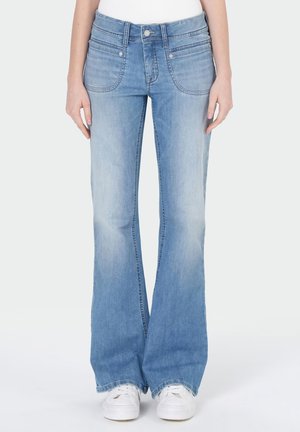 Femme portant un jean flare bleu clair avec des poches plaquées à l'avant et des baskets blanches, debout devant un fond uni.