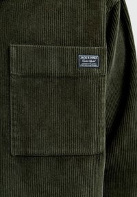 Donkergroene corduroy overhemd met verticale ribbels. Voorzien van een borstzak en een label met de merknaam in witte tekst op een blauwe achtergrond.