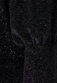 Tecido preto texturizado com brilho metálico, apresentando um design de mangas franzidas e finos toques brilhantes por todo o lado.