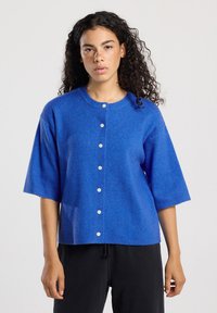 Vrouw met krullend haar en sproeten, gekleed in een blauwe, met knopen gesloten cardigan met mouwen tot de elleboog en zwarte broek, tegen een effen achtergrond.