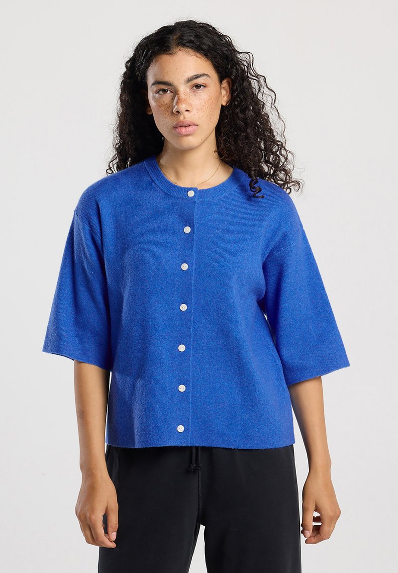 Vrouw met krullend haar en sproeten, gekleed in een blauwe, met knopen gesloten cardigan met mouwen tot de elleboog en zwarte broek, tegen een effen achtergrond.