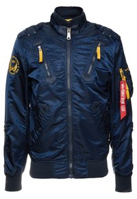 Navy blue bomber jacket made of shiny nylon with multiple zippered pockets, ribbed cuffs, and a red tag labeled "REMOVE BEFORE FLIGHT."