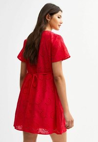 Rotes, kurzärmeliges Kleid mit elastischem Taillengummi, das mit Augenmuster-Stanzungen und einem Bindedetail auf der Rückseite versehen ist. Leichter Stoff, lässige Passform.