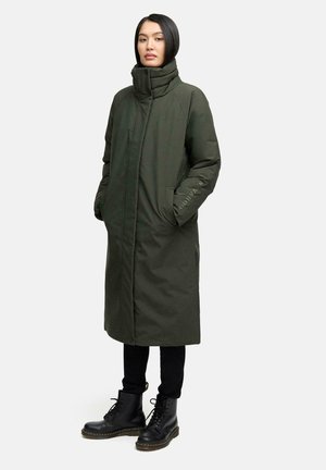 Vrouw staat met een lange donkergroene gewatteerde jas met hoge kraag, zwarte broek en zwarte veterschoenen tegen een effen achtergrond.