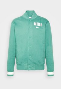 Grön sweatshirt med krage, framknappar och vit "NIKE"-text. Har texturerad tyg och kontrasterande vita detaljer på muddar och nederkant.