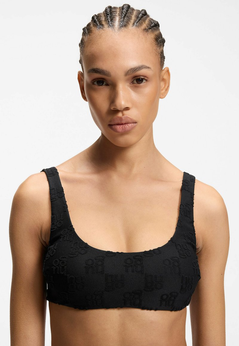 Junge Frau mit geflochtenem Haar, die ein schwarzes, strukturiertes Crop-Top trägt, blickt mit neutralem Gesichtsausdruck nach vorne vor einem schlichten weißen Hintergrund.